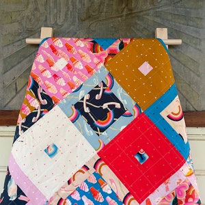 May include: A colourful patchwork quilt featuring a range of patterns, including rainbows, ice cream cones, and stars. The quilt is hanging on a wooden rack against a textured backdrop.