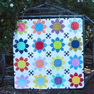 Pode incluir: Um edredom branco com um design de patchwork que apresenta motivos florais coloridos. O edredom é feito de vários tons de azul, vermelho, amarelo, verde e rosa. O edredom está pendurado em uma cerca de madeira.