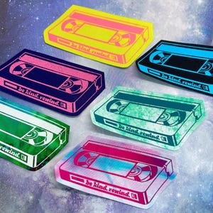 May include: A collection of colorful VHS tape stickers in various hues, including yellow, blue, pink, and green. Each sticker features the text "be kind. rewind." in a retro font, evoking a nostalgic feel. The stickers are arranged on a starry background.