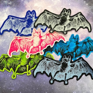 May include: Six bat stickers in various colours, including black, white, blue, green, and pink. The bats are all in a flying position with their wings spread out. The stickers are all on a white background with a galaxy pattern.