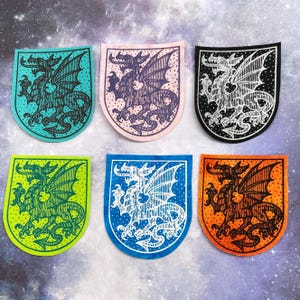 Könnte beinhalten: Sechs farbige Drachenaufnäher in Türkis, Rosa, Schwarz, Limettengrün, Blau und Orange. Jeder Aufnäher zeigt ein detailliertes Drachendesign in Schildform. Die Flügel des Drachen sind ausgebreitet, vor einem Sternenhintergrund.