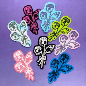 May include: A collection of die-cut floral designs in various colors, including pink, black, blue, and green, with white and black linework. Each design features a bouquet of pansies tied with a bow. The background is a solid purple.