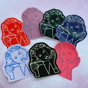 May include: A collection of colorful, die-cut patches featuring a retro illustration of a woman with a finger to her lips. The patches are in various colors, including red, pink, blue, black, and green, with white outlines. The patches are likely for clothing or accessories.