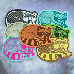May include: A collection of colorful raccoon-shaped stickers. The stickers feature various color schemes, including black and white, teal, orange, green, yellow, and pink. Each sticker has a unique design with a raccoon illustration.