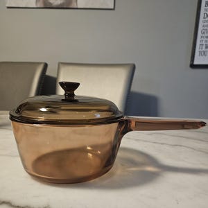 May include: A translucent amber-coloured glass saucepan with a matching glass lid. The round pot features a long, integrated glass handle extending from one side and a small, circular knob centred on the top of the lid for easy removal.