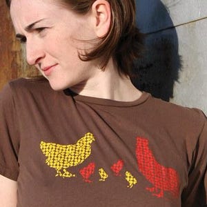 May include: Brown t-shirt featuring a chicken family design. The design includes a yellow chicken, a red chicken, and several smaller red chickens. The shirt is made of a soft material and has short sleeves.