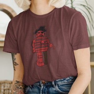 Puede incluir: Una mujer con una camiseta marrón con un robot rojo y negro. El robot lleva un sombrero de pirata y tiene bigote. La mujer está arrodillada sobre una superficie blanca con pintura azul y pinceles.