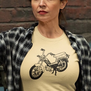 Könnte beinhalten: Eine Frau trägt ein schwarz-weiß kariertes Hemd, ein beiges T-Shirt mit einer schwarz-weißen Illustration eines Motorrads, blaue Jeans und braune Lederhandschuhe. Sie steht in einer Werkstatt mit einem Motorrad im Hintergrund.