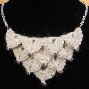 May include: A cream-colored crochet necklace with a silver chain. The necklace features a layered, scalloped design with small, dark purple beads. The crochet work has a textured appearance, and the necklace is displayed against a black background.