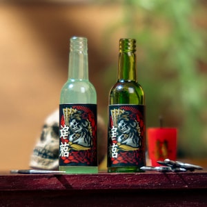 May include: Two small glass bottles, one green and one clear, with black labels featuring a skull design and Japanese text. The bottles are on a wooden surface, with a skull and red candle in the background.