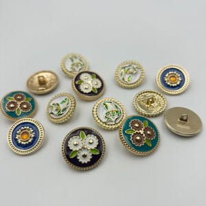 Puede incluir: Una colección de botones decorativos con diseños florales. Los botones presentan bordes y respaldos de metal dorado, con diseños de esmalte coloridos en verde azulado, azul marino, blanco y dorado. Algunos botones tienen un patrón floral en relieve.