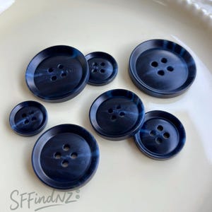 Puede incluir: Un conjunto de botones azul marino con efecto veteado. Los botones, de diferentes tamaños, tienen cuatro agujeros. Se muestran sobre una superficie crema. El texto "SFfindNZ" es visible en la esquina inferior izquierda.