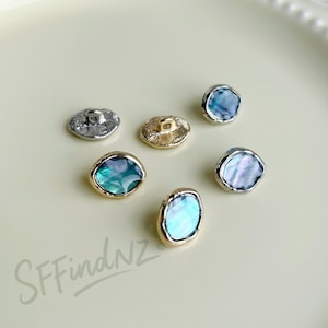 Può includere: Una collezione di bottoni in conchiglia iridescente di varie forme. I bottoni hanno un bordo metallico in tonalità argento e oro. Le conchiglie mostrano sfumature di blu, verde e viola. I bottoni sono disposti su una superficie chiara.