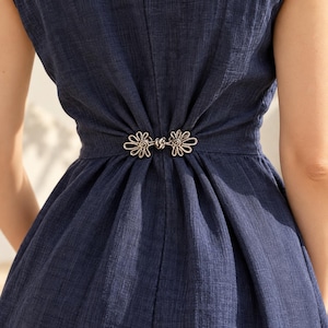 May include: A dark blue sleeveless dress featuring a textured fabric and a central silver-toned metal clasp at the waist. The clasp has a decorative floral-inspired shape with intricate wire detailing, pulling the fabric into soft gathers.