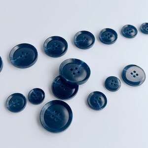 May include: A collection of navy blue buttons in various sizes, arranged on a white surface. Each button has four holes and a marbled appearance, creating a subtle texture. The buttons range from small to large.