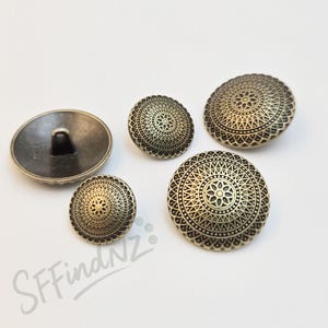 May include: Five antique brass-coloured buttons with intricate, circular, embossed designs. The buttons vary in size, with one showing the back with a shank. The buttons are arranged on a white surface.