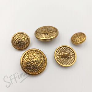 May include: A collection of five gold-colored buttons. The buttons feature embossed designs, including a shield with a horse and other symbols. The buttons vary in size and have a textured surface.