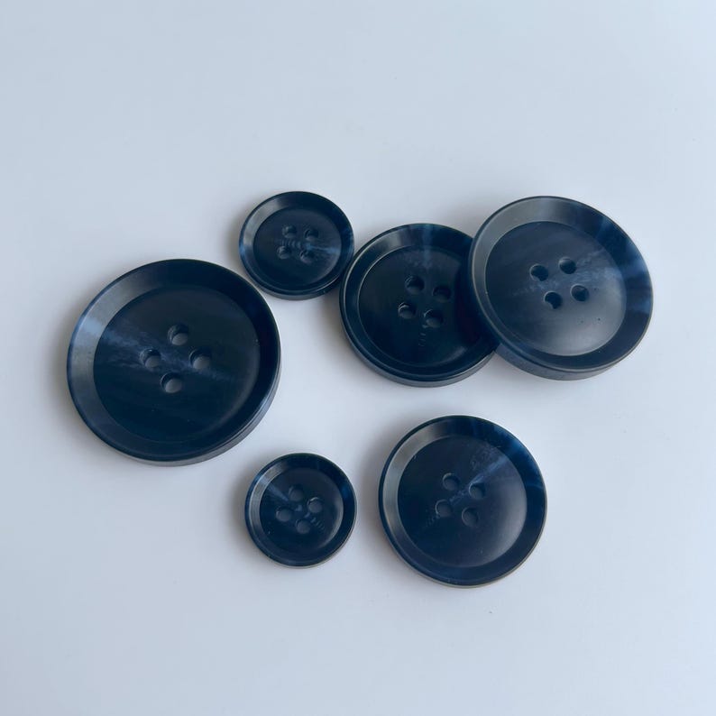 May include: A collection of navy blue buttons in various sizes. Each button has four holes and a subtle marbled effect. The buttons are arranged on a white surface, showcasing their different diameters.
