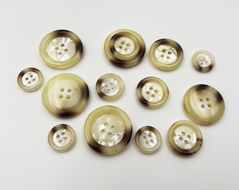 May include: A collection of various-sized buttons in shades of cream and brown. The buttons have four holes and a pearlescent center. The outer edges of the buttons have a darker brown ring. The buttons are arranged on a white background.