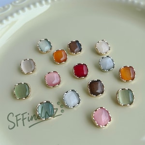 Puede incluir: Una colección de cuentas de cabujón cuadradas y coloridas con bordes de metal dorado. Las cuentas son de varios colores, incluyendo rosa, verde, naranja, rojo y marrón. Las cuentas están dispersas sobre una superficie de color amarillo claro. El texto "SFFindNZ" está en la esquina inferior izquierda.