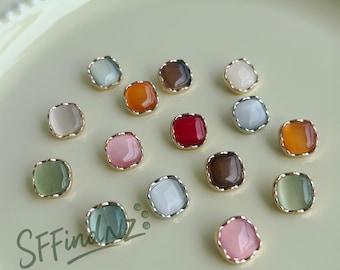 May include: A collection of square-shaped, colorful cabochon beads with gold-colored metal edges. The beads are in various colors, including pink, green, orange, red, and brown. The beads are scattered on a light yellow surface. The text "SFFindNZ" is in the lower left corner.