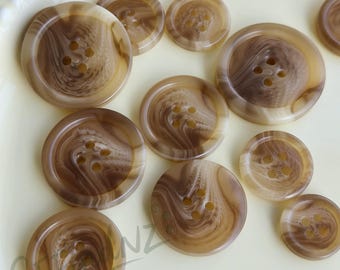 May include: A collection of brown and tan swirl-patterned buttons in various sizes. The buttons have four holes for sewing and a glossy finish. The buttons are arranged on a light yellow surface.
