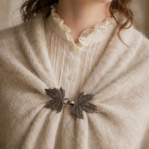Può includere: Una fibbia in metallo color argento con due dettagliate forme di foglie d'acero collegate da un anello centrale. La fibbia è fissata a uno scialle in maglia beige, testurizzato e soffice, indossato sopra una camicia color crema con colletto in pizzo.