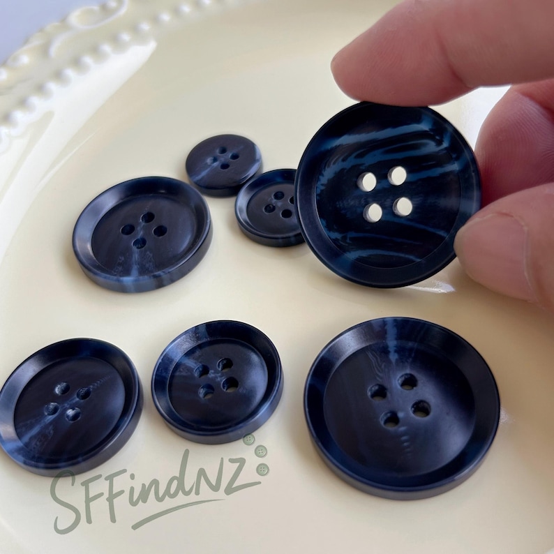 May include: A collection of navy blue buttons with a marbled effect and four holes. The buttons vary in size, with one being held by a hand. The buttons are on a cream-colored surface. The text "SFfindNZ" is visible.