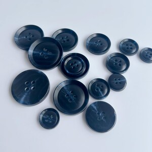 May include: A collection of navy blue buttons in various sizes. The buttons have a marbled appearance with four holes each. The buttons are arranged on a white surface, showcasing their different diameters and designs.