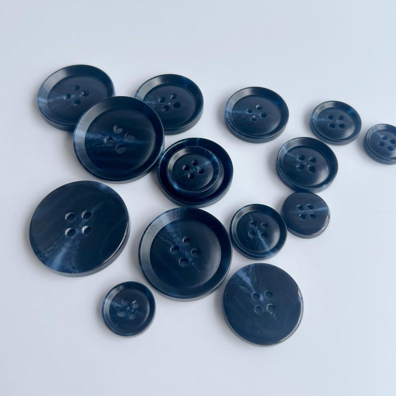 May include: A collection of navy blue buttons in various sizes. The buttons have a marbled appearance with four holes each. The buttons are arranged on a white surface, showcasing their different diameters and designs.