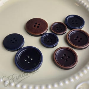 Puede incluir: Una colección de botones de varios tamaños en tonos marrón y azul marino. Los botones tienen cuatro agujeros y están dispuestos sobre un plato amarillo claro. Algunos botones tienen un borde contrastante.
