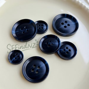 May include: A collection of navy blue buttons with a marbled effect. The buttons vary in size and have four holes each. The buttons are arranged on a cream-colored surface. The text "SFindNZ" is visible.