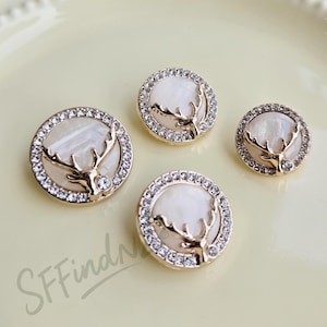 May include: Four round decorative buttons with a gold-tone rim and a white center. Each button features a gold-tone deer antler design and is encrusted with small, clear rhinestones. The buttons are set against a light yellow background.