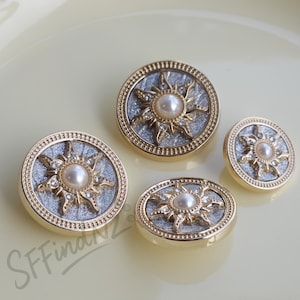 Könnte beinhalten: Vier dekorative Knöpfe mit einem Sonnenmuster. Die Knöpfe haben einen goldfarbenen Rand, eine silberfarbene Mitte und eine Perle in der Mitte. Der Sonnenstrahl ist goldfarben mit kleinen, klaren Kristallen. Ein Knopf ist oval.