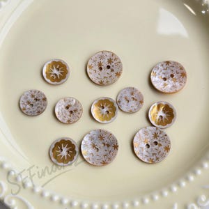 Pode incluir: Uma coleção de botões decorativos em vários tamanhos. Os botões apresentam uma base branca com desenhos florais e de estrelas douradas. Cada botão tem dois furos para costurar.