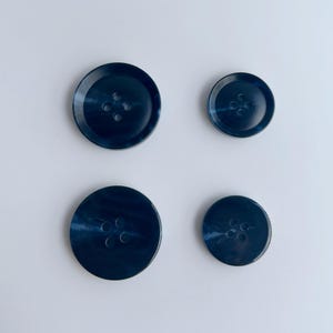 May include: Four navy blue buttons of varying sizes, each with four holes. The buttons have a marbled appearance, with lighter blue swirls throughout. The buttons are arranged on a white background.