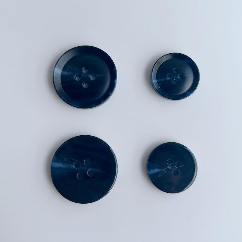 May include: Four navy blue buttons of varying sizes, each with four holes. The buttons have a marbled appearance, with lighter blue swirls throughout. The buttons are arranged on a white background.