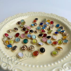 Peut inclure: Un assortiment de boutons ronds colorés avec des bordures dorées. Les boutons présentent diverses couleurs, notamment rouge, vert, bleu, jaune et rose. Certains boutons ont un aspect texturé, semblable à une gemme. Les boutons sont disposés sur une assiette décorative jaune clair.