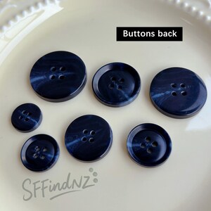 May include: A collection of seven navy blue buttons with a marbled effect. The buttons vary in size and have four holes each. The text "Buttons back" is displayed in a black rectangle. The buttons are arranged on a white surface.