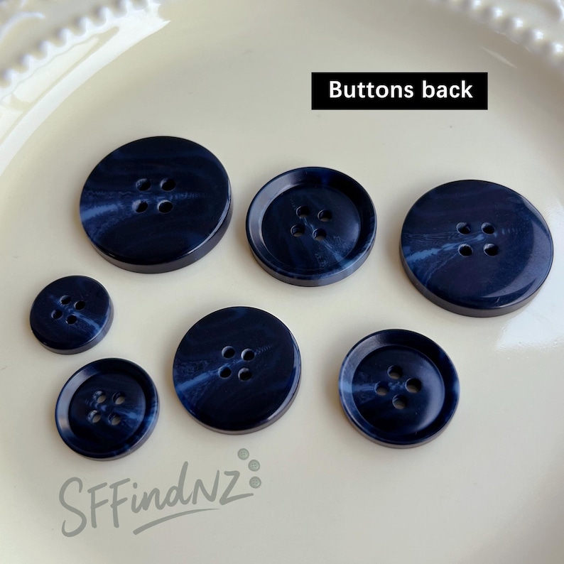 May include: A collection of seven navy blue buttons with a marbled effect. The buttons vary in size and have four holes each. The text "Buttons back" is displayed in a black rectangle. The buttons are arranged on a white surface.