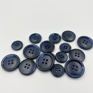 May include: A collection of navy blue buttons in various sizes. Each button has four holes and a slightly marbled appearance. The buttons are arranged on a white surface, showcasing their different diameters and designs.