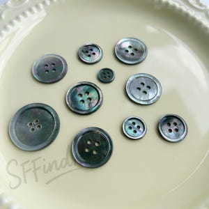 May include: A collection of iridescent, dark gray buttons of varying sizes. The buttons have a pearlescent sheen and feature two or four holes for sewing. The buttons are arranged on a light yellow, scalloped-edge plate.