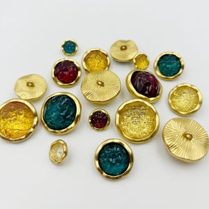 Könnte beinhalten: Eine Sammlung runder Knöpfe in verschiedenen Größen mit goldfarbenen Metallrändern. Einige Knöpfe haben strukturierte goldene Mitten mit radialen Linien, während andere durchscheinende, farbige Einsätze in Rot, Gelb und Blaugrün aufweisen.