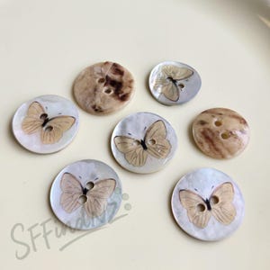 Puede incluir: Una colección de botones redondos con diseños de mariposas. Los botones presentan una mezcla de colores, incluyendo beige, marrón y blanco. Algunos botones tienen un acabado nacarado, mientras que otros tienen una apariencia veteada. Cada botón tiene dos agujeros para coser.