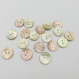 Könnte beinhalten: Eine Sammlung von neunzehn runden Knöpfen mit perlmuttartigem Glanz in schillernden Tönen von Blassgrün, zartem Rosa und Gold. Jeder Knopf hat zwei mittige Löcher und ein feines, kompliziertes eingraviertes Muster auf der Oberfläche.