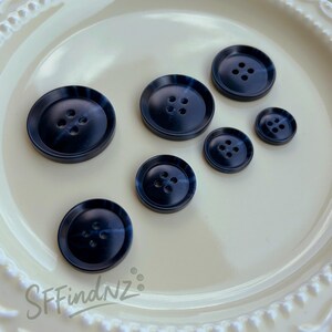 May include: A collection of navy blue buttons in various sizes, each with four holes. The buttons have a marbled effect and are arranged on a cream-colored plate. The text "SFFindNZ" is visible in the lower left corner.
