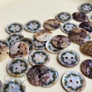 May include: A collection of round, two-hole buttons made from iridescent shell material. Some buttons feature a printed green and yellow floral wreath pattern, while others display a natural, mottled brown and cream marbled finish.