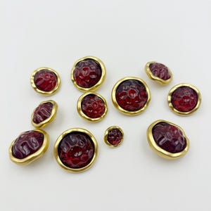Puede incluir: Conjunto de botones rojos texturizados con bordes dorados. Los botones, de diferentes tamaños, están dispersos sobre una superficie blanca. Los centros rojos presentan una textura burbujeante única, agregando interés visual.
