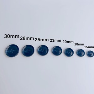 May include: A collection of seven navy blue buttons of varying sizes, ranging from 15mm to 30mm in diameter. Each button features four holes for sewing and a slightly domed surface. The buttons are arranged in a row, showcasing their size differences.