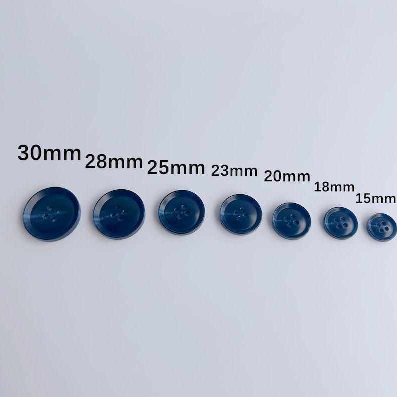 May include: A collection of seven navy blue buttons of varying sizes, ranging from 15mm to 30mm in diameter. Each button features four holes for sewing and a slightly domed surface. The buttons are arranged in a row, showcasing their size differences.